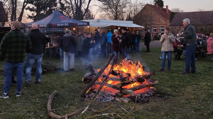 Menschen stehen in der Dämmerung um ein Lagerfeuer. Im Hintergrund befinden sich Imbiss-Zelte.
