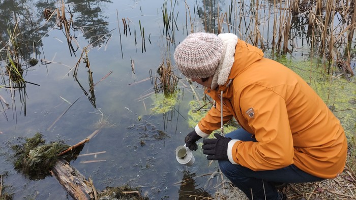 Eine Wanderin schöpft Wasser aus einem Teich. Es ist früher Morgen und der Nebel hängt noch über dem Wasser.