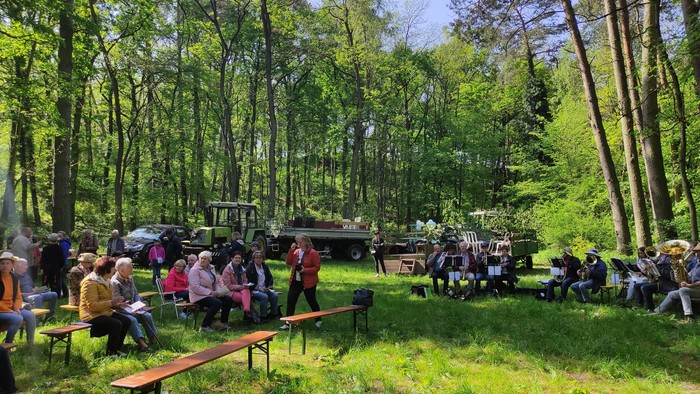 Auf einer Waldlichtung sitzen Menschen auf Bänken. Etwas abseits sitzt der Posaunenchor. Im Hintergrund Traktoren mit Anhängern zum Sitzen.