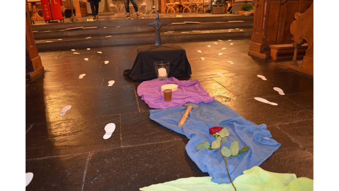Künstlerische Bodeninstallation mit einem Kreuz, Tuch, Rose und verstreuten Gegenständen in einer Kirchenumgebung.