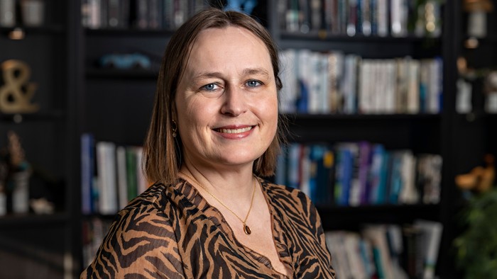 En smilende kvinde i en zebrastribet top står i et hyggeligt, velassorteret rum med bogreoler.