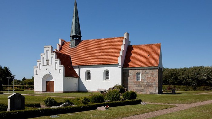En hvid kirke med rødt tag og spir med tårn, omgivet af græs og træer