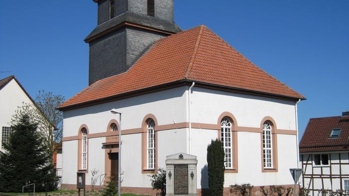 Außenansicht der evangelischen Kirche in Niederlistingen. Das Gebäude hat eine helle Fassade mit hohen Rundbogenfenstern und ein rotes Ziegeldach. Der quadratische Turm ist im oberen Bereich mit dunklen Schieferplatten verkleidet und trägt ein kleines spi