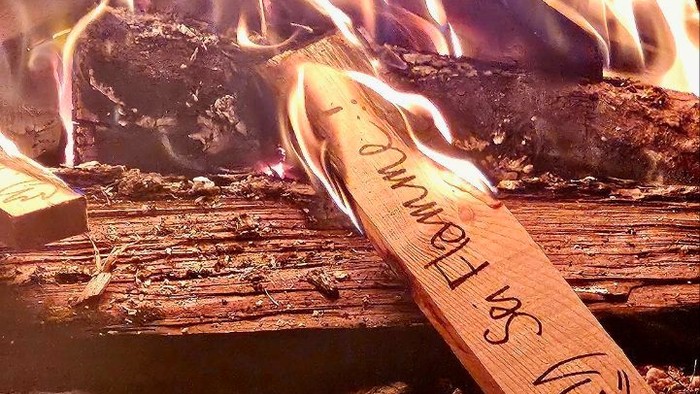 Flamme eines Lagerfeuers, darin ein Holzschild mit Aufschrift 