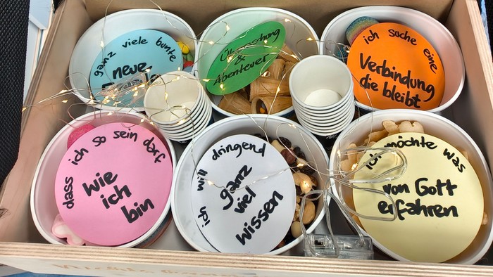 In einem Bauchladen sind sechs verschiedene Becher mit Süßigkeiten, darauf liegen runde Karten, die mit Kategorien beschriftet sind