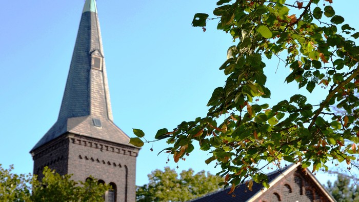 Kirchturm mit Kreuz und goldener Spitze, umgeben von Bäumen und grünem Blattwerk