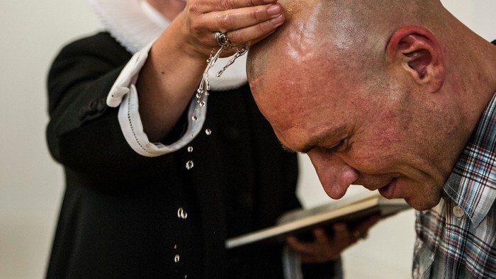 En bald mand læser fra en bog mens en anden person holder en rosary ved hans hoved.