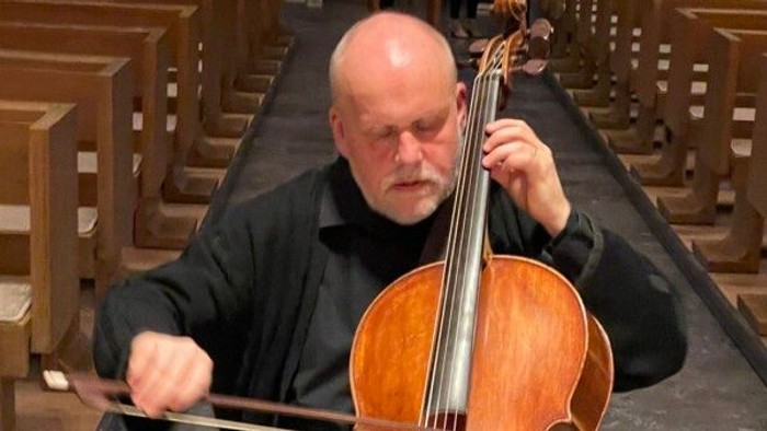 Ein glatzköpfiger Mann spielt ein Cello in einer Kirche mit hölzernen Kirchenbänken und einem Holzboden.