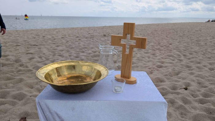 An einem Strandabschnitt der Ostsee steht ein kleiner Tisch mit einer weißen Tischdecke. Darauf sind eine Taufschale aus Messing und ein Holzkreuz platziert. Außerdem ein Krug mit Wasser und ein Glas mit einer Kerze darin.