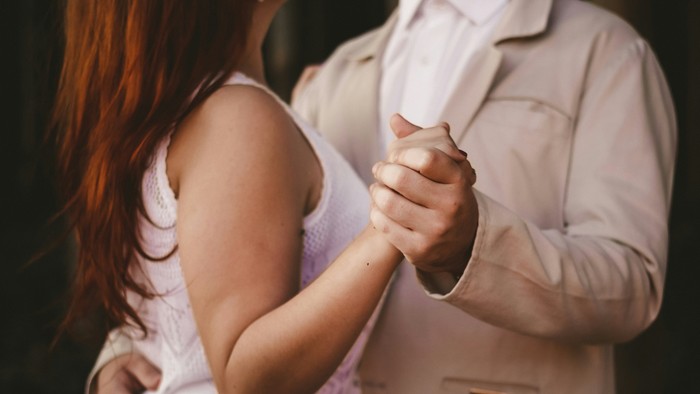 Ein Paar hält sich an den Händen, die Frau hat rotes Haar und trägt ein weißes Kleid, der Mann ein hellfarbiges Hemd.