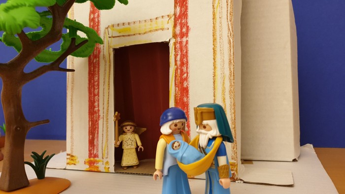 Spielzeugfiguren in einer Weihnachtskrippe, die in der Nähe eines geschmückten Türrahmens mit einem Baum draußen stehen.