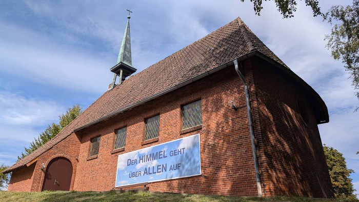 Das Bild zeigt eine rote Backsteinkirche mit einem kleinen Turm und einem Banner mit der Aufschrift
