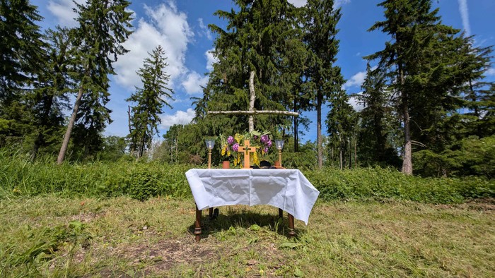 Auf einer Lichtung im Wald steht ein mit Blumen geschmückter Altar, dahinter ein Kreuz aus Birken.