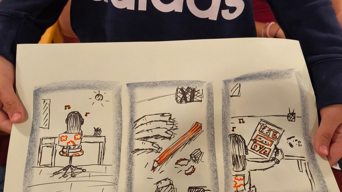 Eine jugendliche Person hält ein selbstgemaltes Bild in die Kamera, es zeigt wie im Comic drei Kästchen, darin abgebildet ist ein Mädchen, das am Schreibtisch etwas malt.