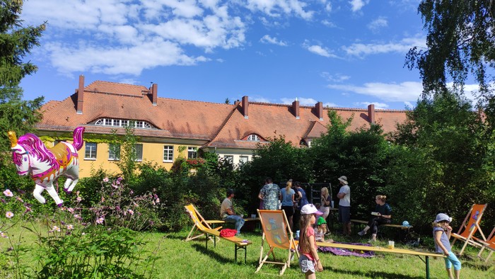 In einem grünen Garten sind Kinder und Erwachsene, auf der linken Seite schwebt ein Einhornheliumballon
