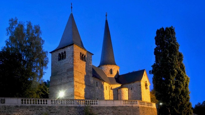 Eine wunderschön beleuchtete historische Kirche mit Zwillingstürmen erhebt sich vor dem Abendhimmel, umgeben von einer Steinmauer.