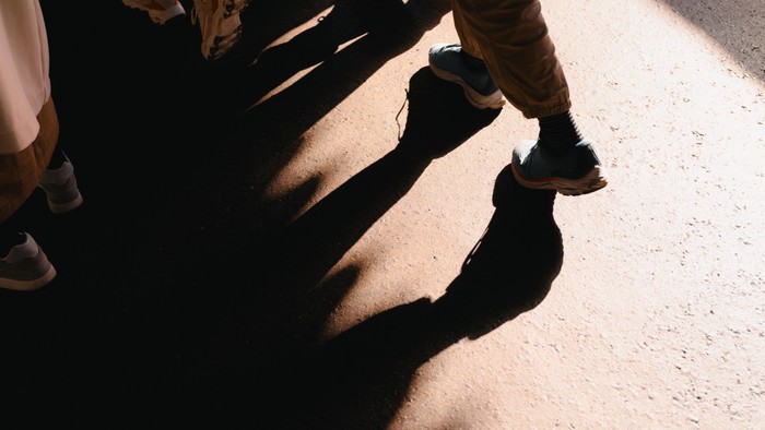 Schatten von Beinen und Schuhen auf sonnenbeschienenem Gehweg.
