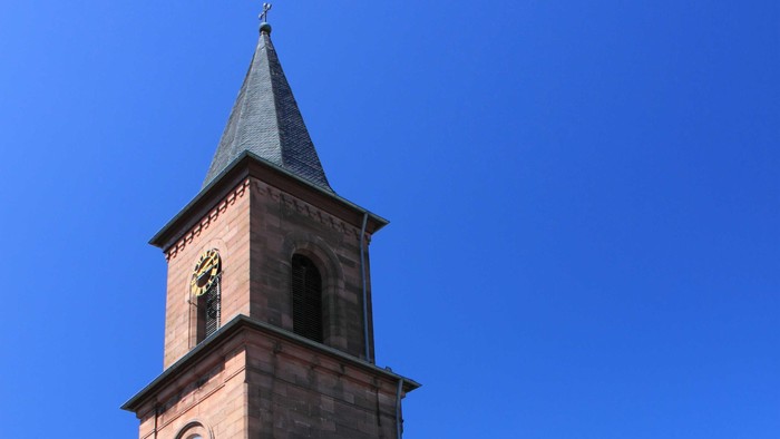 Kirche mit hohem Uhrturm und Spitzhelm vor klarem blauen Himmel.