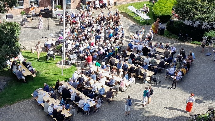 Stor udendørs forsamling med mennesker siddende på bænke og stående, sandsynligvis til en dimissionsceremoni.