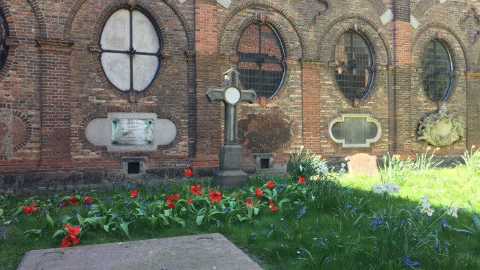 Ein ruhiges Grab, geschmückt mit Blumen und einem Kreuz, vor einer Ziegelmauer mit Bogenfenstern.