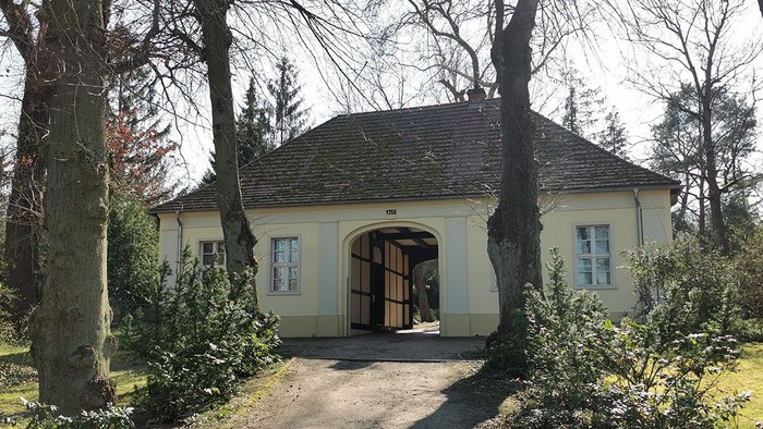 Ein kleines, gelbes Gebäude mit einem Torbogen, umgeben von Bäumen und Büschen.