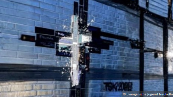 Eine geziegelte Wand, an der einzelne Ziegel schwarz gefärbt sind und ein Jesus Kreuz darstellen. Auf das Kreuz selbst ist ein Jesus Kreuz in weiß als Graffiti projiziert. 
