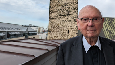 Ein älterer Mann in formeller Kleidung steht auf einem Dach und blickt auf historische Backsteingebäude hinab.