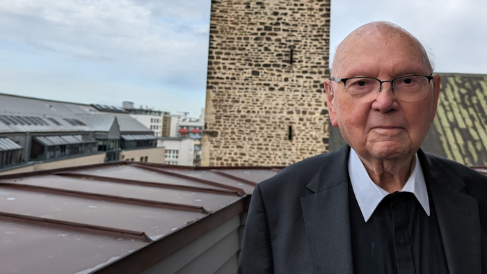 Ein älterer Mann in formeller Kleidung steht auf einem Dach und blickt auf historische Backsteingebäude hinab.