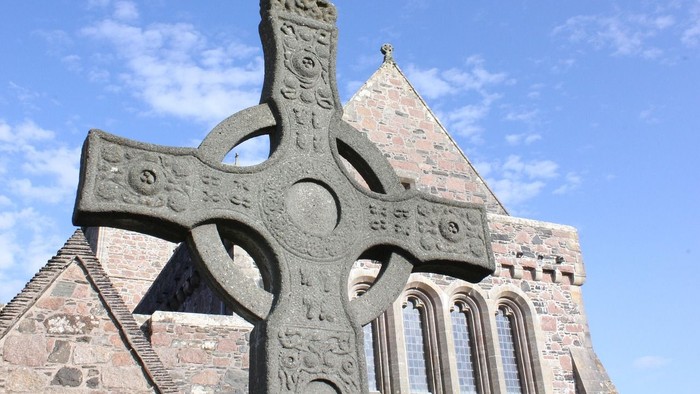 Hochdetailliertes keltisches Kreuz vor einer alten Steinkirche unter blauem Himmel.