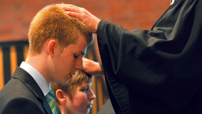 Ein Mann in schwarzer Robe berührt den Kopf eines jungen Mannes in Anzug.
