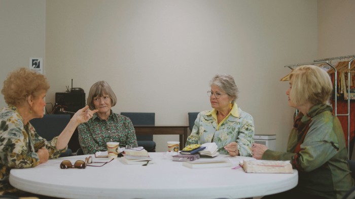 Vier Frauen sitzen um einen Tisch in einem Meeting und unterhalten sich, mit Papieren und Tassen vor sich.