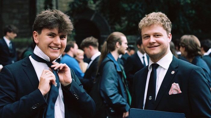 Vier junge Männer in formeller Kleidung, die bei einer Abschlussfeier oder einem offiziellen Anlass lächeln.