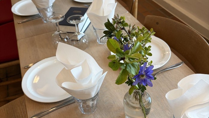 Elegant opstillet spisebord med tallerkener, servietter, blomster og vandglas på træstole.