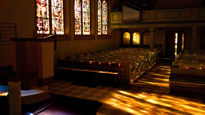 Das Bild zeigt das Innere einer Kirche mit Blick in den Kirchenraum. Links steht eine Kanzel mit Lesepult, davor liegt ein aufgeschlagenes Buch. Reihen von hellen Kirchenbänken erstrecken sich nach hinten durch den Raum. An der linken Wand befinden sich 