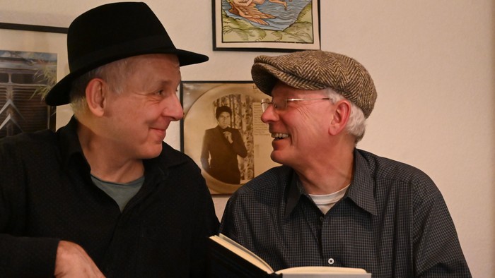 Zwei lächelnde Männer mit Hüten teilen einen heiteren Moment, während sie gemeinsam ein Buch halten.