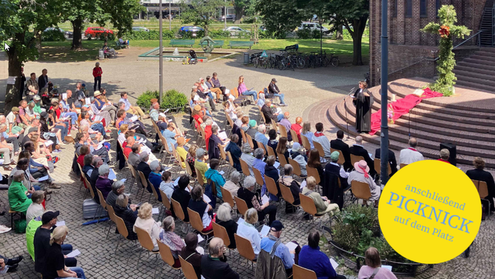 Große Menschenmenge sitzt im Freien bei einem Picknick, viele halten gelbe Plakate
