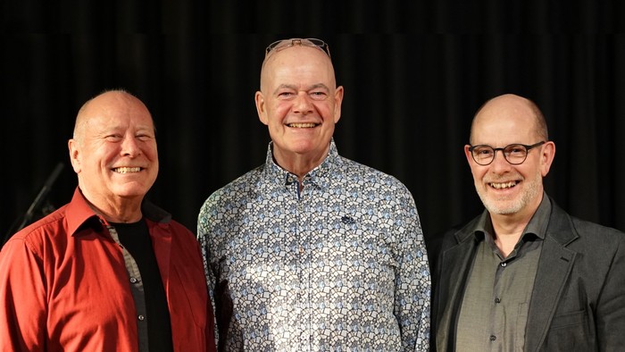 Tre smilende mænd i formelt tøj poser sammen på en scene.