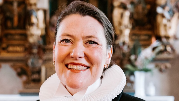 En kvinde i sort jakke med hvid krage står foran en prægtig altertavle.