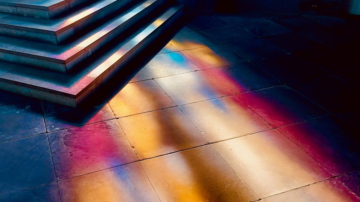 Lebendige Lichtstreifen erzeugen eine farbenfrohe Reflexion auf einem dunklen, gefliesten Boden unter einer Treppe.