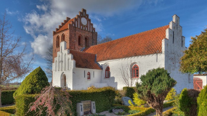 Et hvidt kirkebygning med røde tag og en klokketårn, omgivet af grønne buske og træer under et blåt himmel.