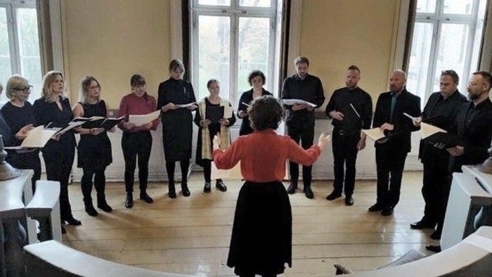 En gruppe af mænd og kvinder synger i en sangskole under ledelse af en instruktør.