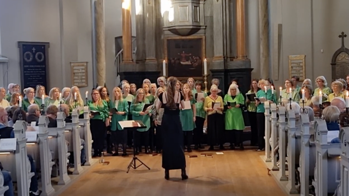 Et stort kor i grønne kjortler optræder i en historisk kirke med et publikum siddende på træbænke.