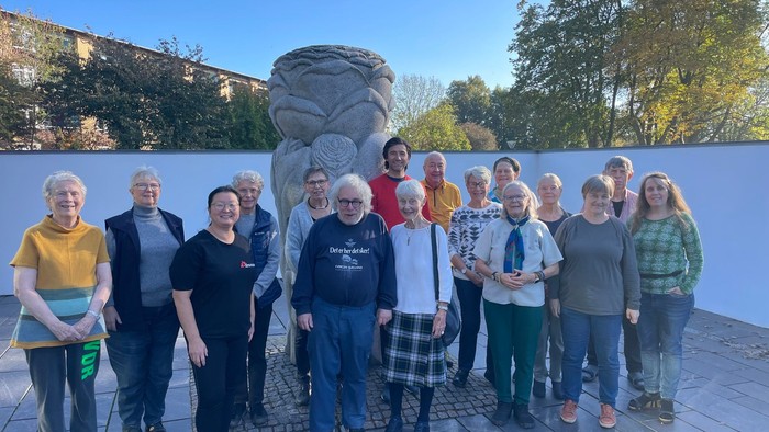 En gruppe af ældre personer står sammen ved en stor skulptur i en park