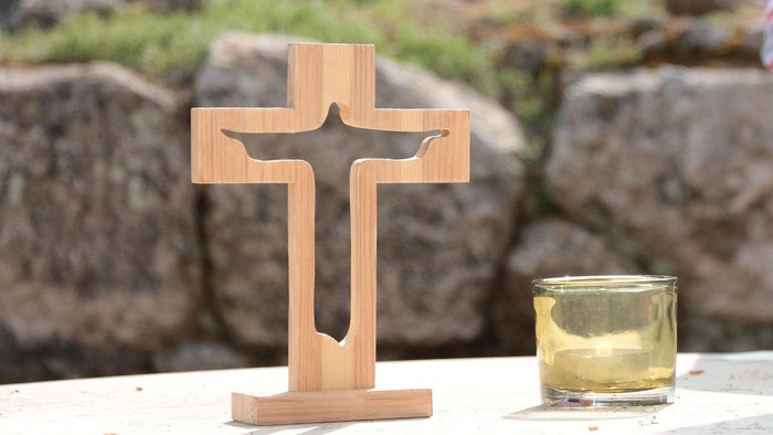 Holzkreuz mit einer Kerze in einem kleinen Glasbehälter auf einer Steinfläche im Freien.