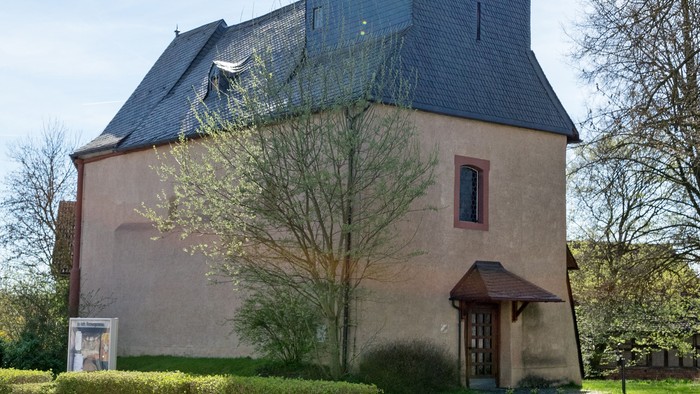 Kirche mit hohem Turm und Kreuz, umgeben von Bäumen und Gras.