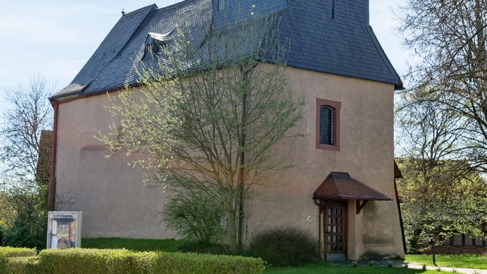Kirche mit hohem Turm und Kreuz, umgeben von Bäumen und Gras.