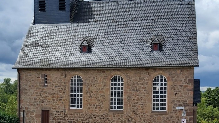 Steinerne Kirche mit hohem Glockenturm und steilem Dach, umgeben von Bäumen.