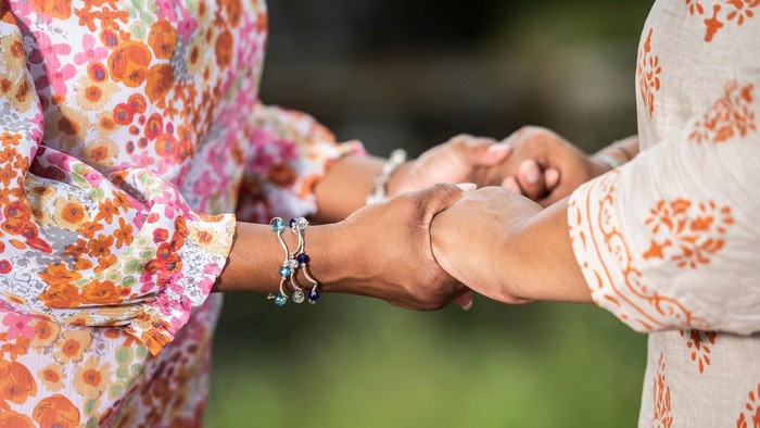 Zwei Personen halten sich draußen an den Händen, tragen blumige und gemusterte Kleidung mit Armbändern.
