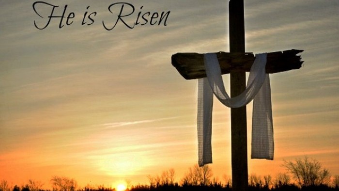 Cross with white cloth at sunset, "He is Risen" written above