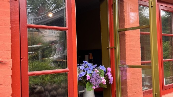 Helles rotes Backsteinhaus mit offenen, grün gerahmten Fenstern, die violette Blumen und die Aussicht nach draußen zeigen.
